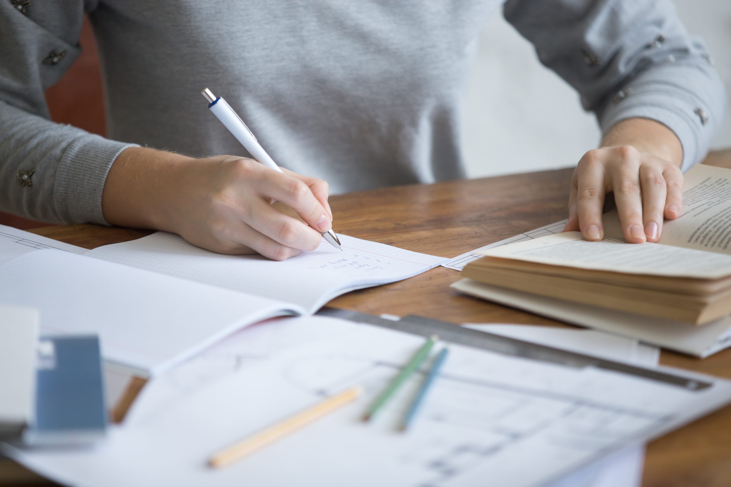 Person writing notes in a notebook with books and papers on a desk.