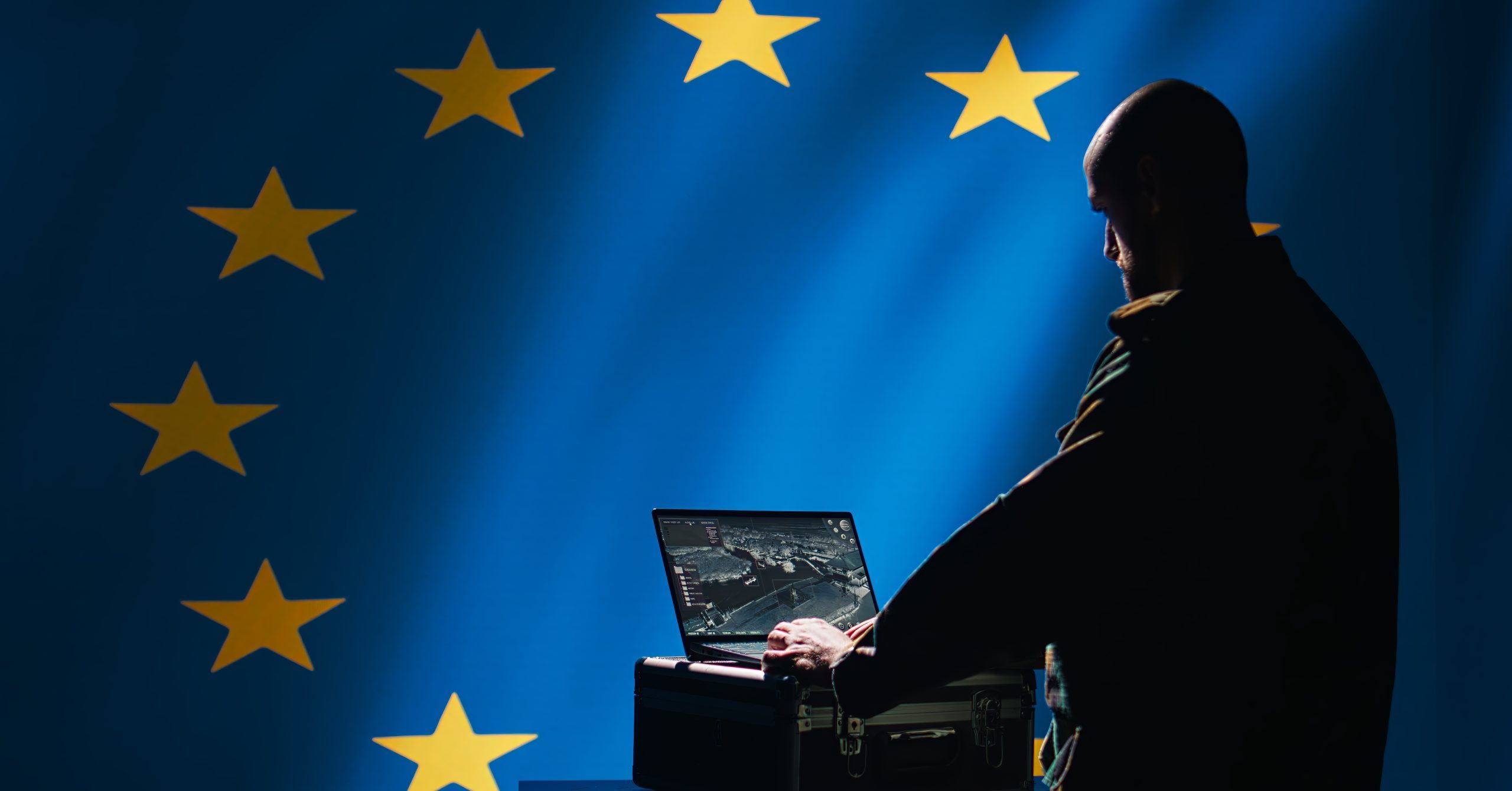 Person using a laptop in front of an EU flag, suggesting cyber or intelligence activity