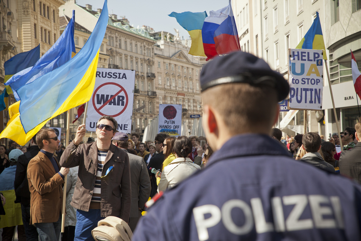 A street protest in a European city where a crowd holds Ukrainian flags and signs opposing Russia’s war.