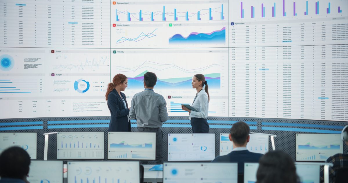 Team of data scientists discussing graphs and reports on big digital screen in monitoring office.