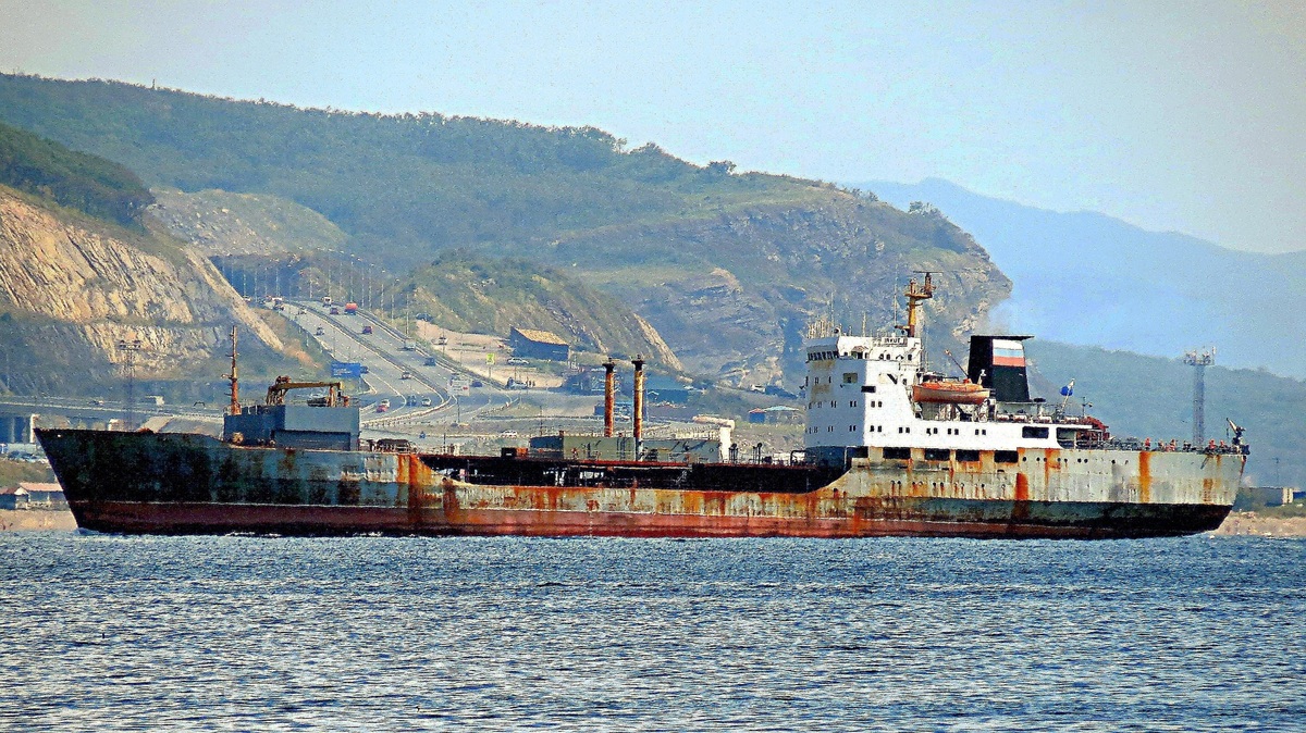 Rusted oil tanker with Russian flag on the sea. Behind the ship is a hill and a highway with cars.