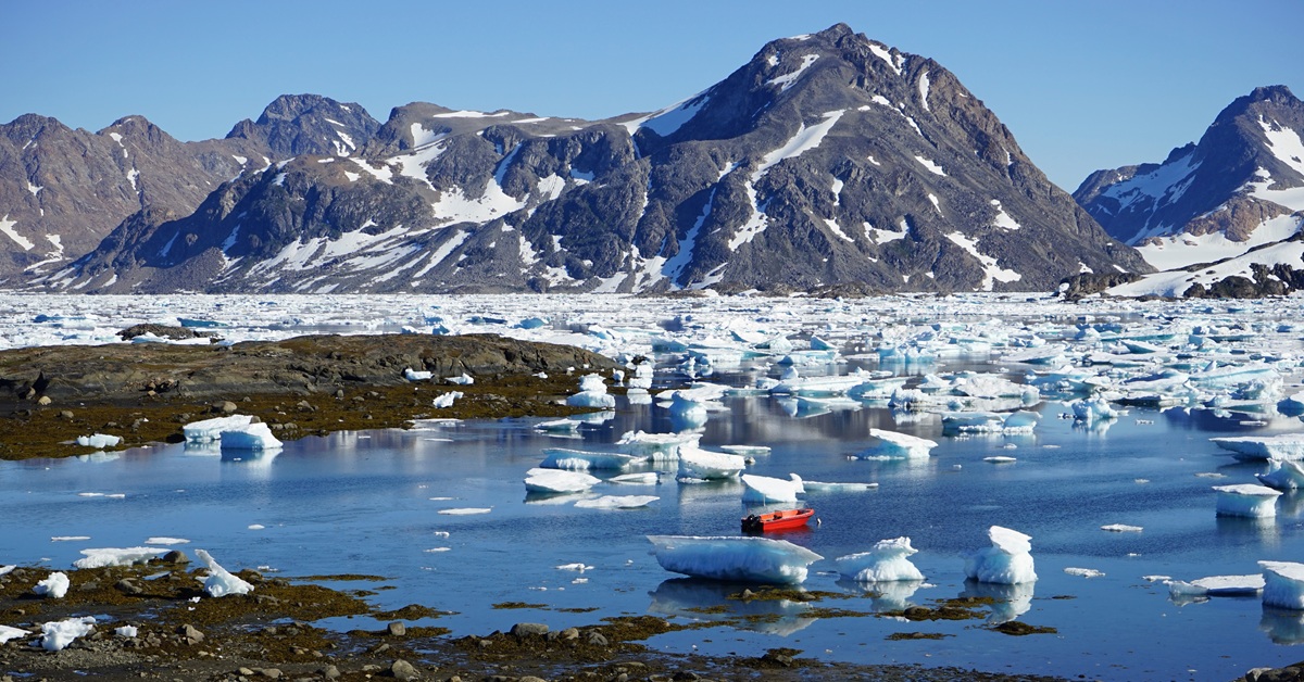 Arctic coastal landscape with rugged snow-capped mountains rising behind a calm bay scattered with floating icebergs and sea ice. A small red boat rests among the ice, creating a striking contrast against the blue water and white ice under a clear sky.