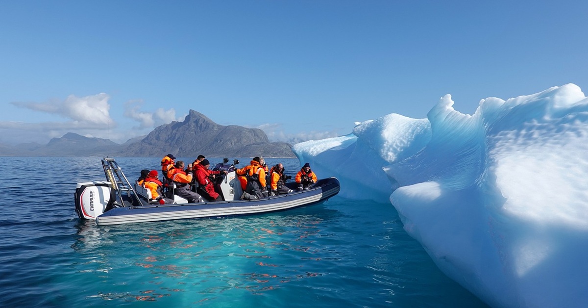 A group of people wearing bright orange cold-weather survival suits travel in a rigid inflatable boat alongside a large blue-white iceberg in calm Arctic waters.