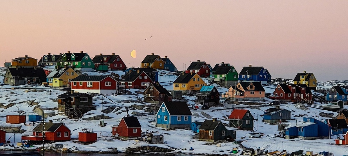 Colorful wooden houses in shades of blue, red, yellow, green, and pink are scattered across a snow-covered rocky hillside in a Greenlandic coastal settlement. The pale moon hangs low above the horizon under a soft pink evening sky, while small boats and sheds line the waterfront in the foreground.