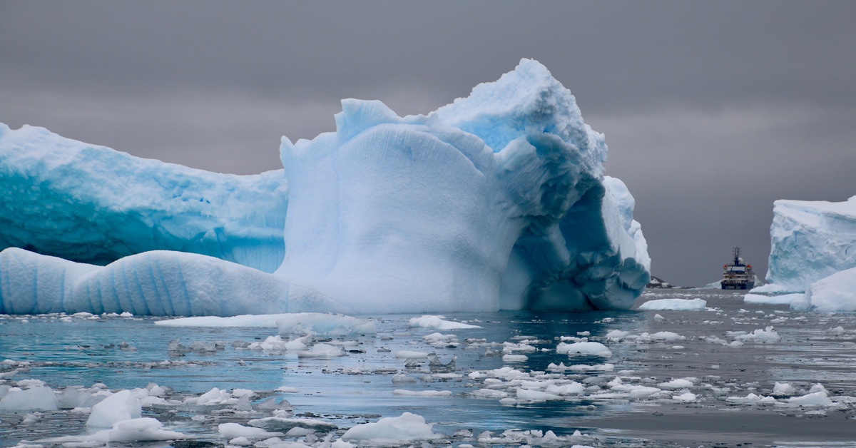 A large, irregular iceberg is floating in the sea. In the distance, a vessel can be seen sailing away.
