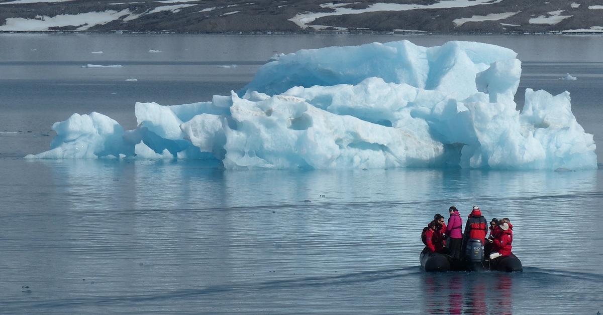 A rubber boat filled with people is heading toward an iceberg floating in the sea. In the background, a snow-covered shoreline is visible.