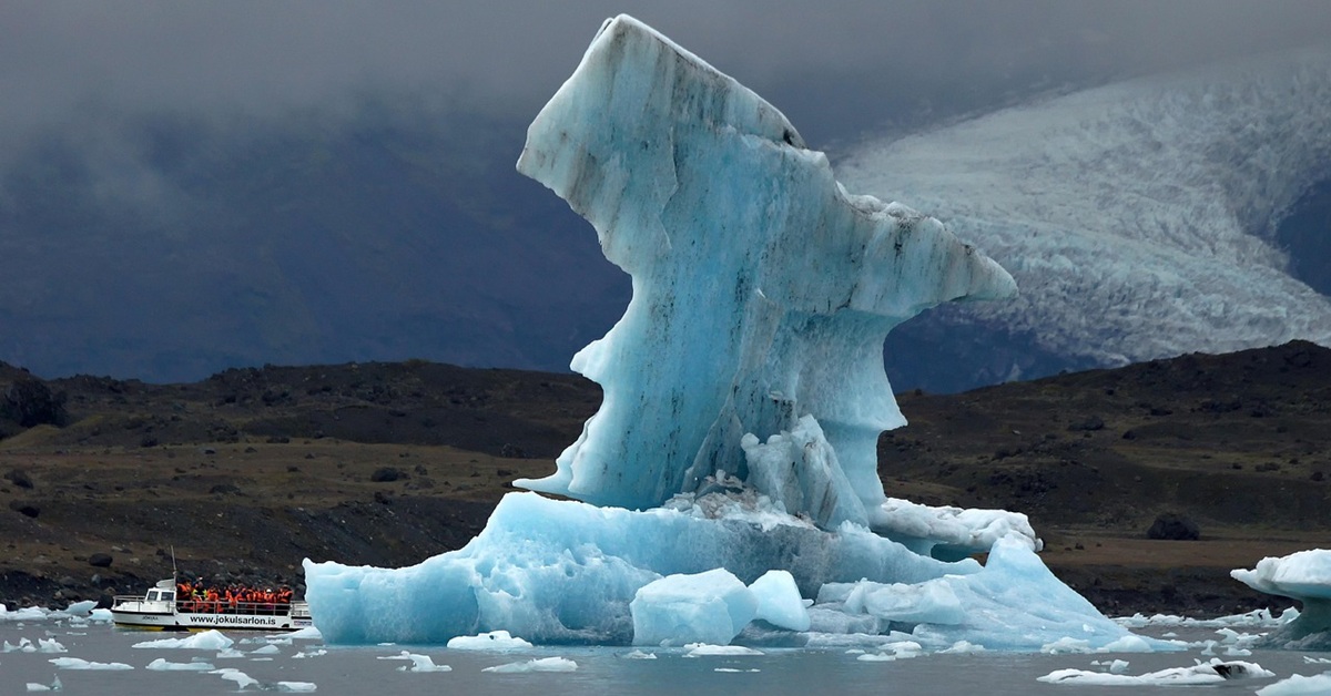 A tall chunk that has broken off from an iceberg floats in the sea. Beside it is a tourist boat, and in the background a shoreline and glacier are visible.