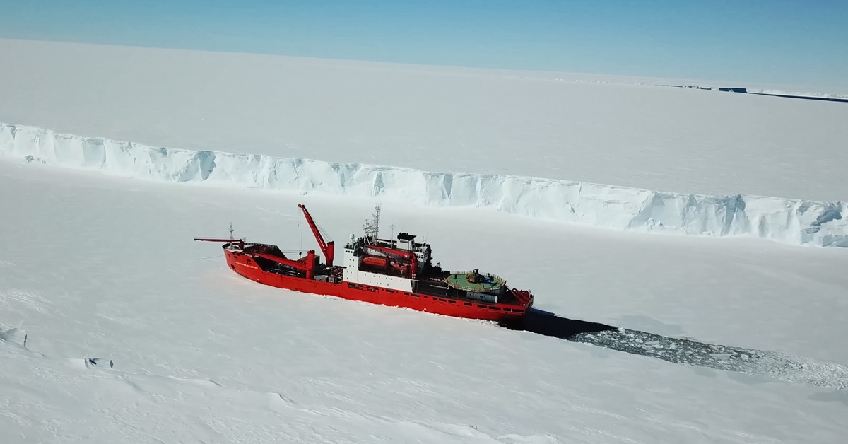 A red-and-white icebreaker is opening a navigation channel in the middle of a white snow and ice landscape.