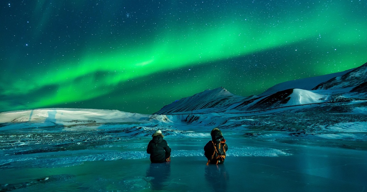 Green northern lights in the sky, snow-capped mountains in the background, and two people sitting on a glacier in the foreground watching