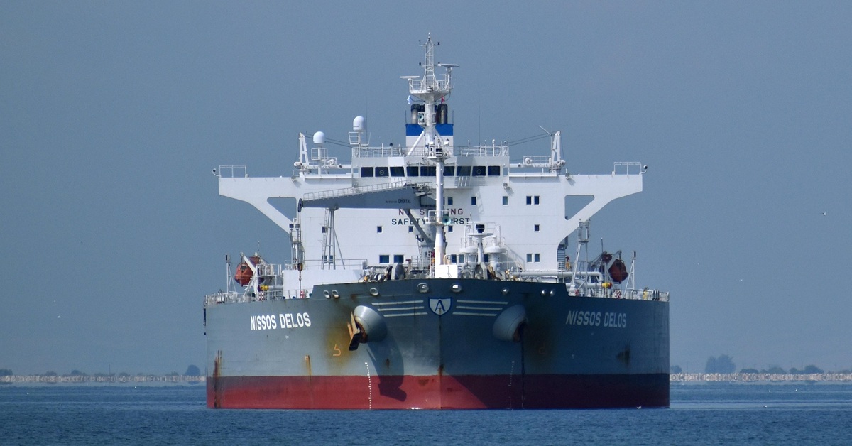 Front view of a large oil tanker sailing on calm water under a hazy blue-grey sky. The vessel dominates the frame, with its massive bow, white superstructure, and navigation equipment clearly visible. A low shoreline stretches across the distant horizon.