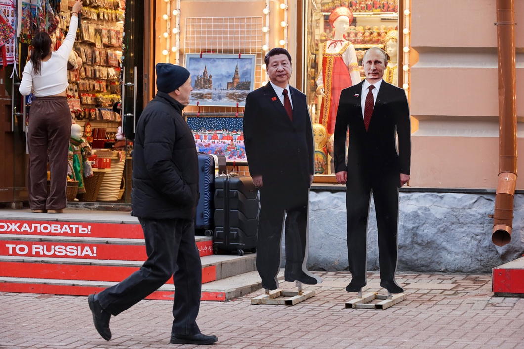 Life-size cardboard cutouts of Xi Jinping and Vladimir Putin stand outside a Russian souvenir shop, while a passer-by walks past them. Russian-themed souvenirs, dolls and artwork are displayed in the shop window.