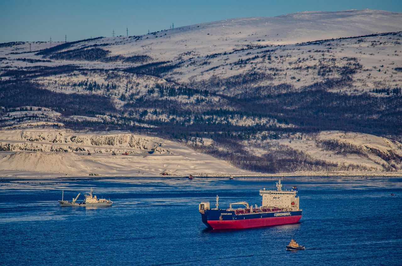 A cargo ship sails through the blue waters of Murmansk, Russia, with a small tugboat nearby and snow-covered hills in the background.